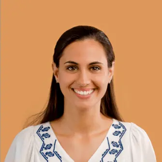 digital marketing company in kuwait-Smiling woman with long dark hair wearing a white blouse with blue embroidery, standing against a warm beige background